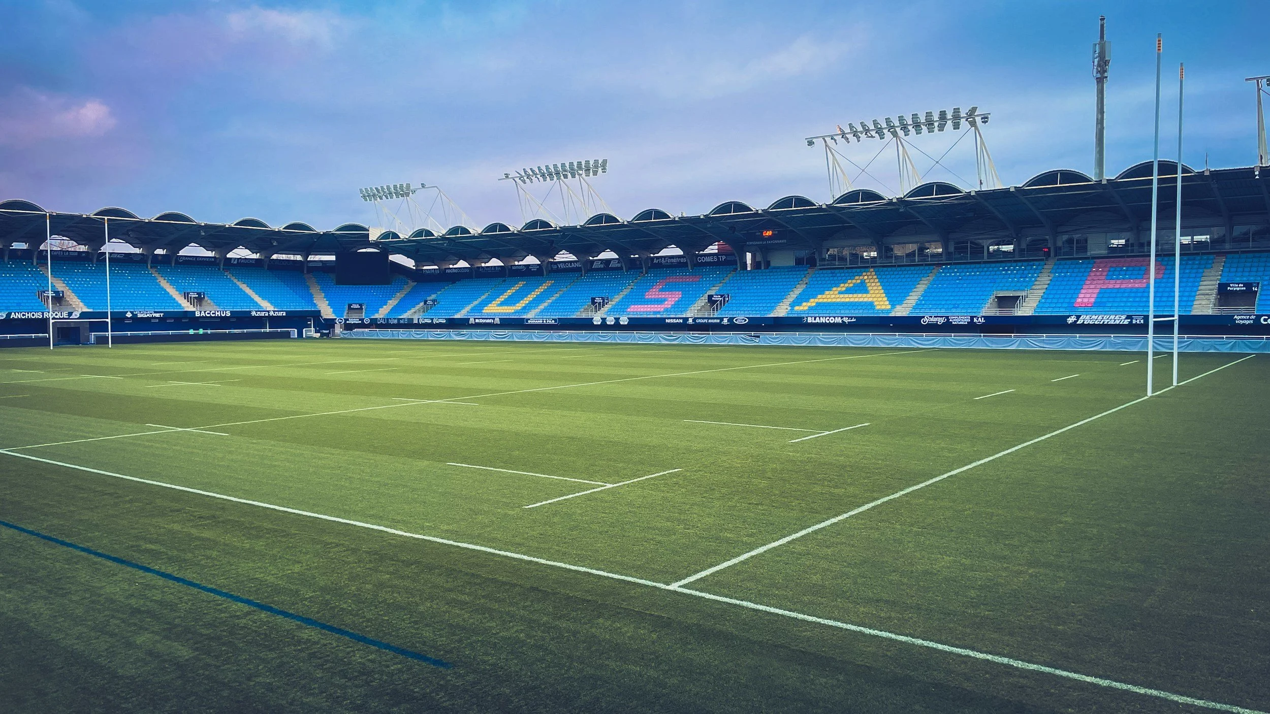 Empty rugby field inside a stadium with blue seating and colorful letters spelling 'OLYMPIQUE' on the stands, under a clear sky.