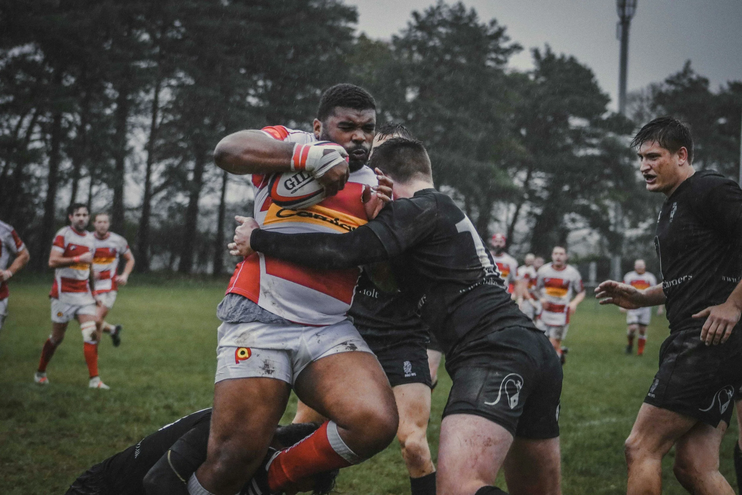 Rugby match with players in action, one player carrying the ball while others attempt to tackle him, on a grassy field with trees in background
