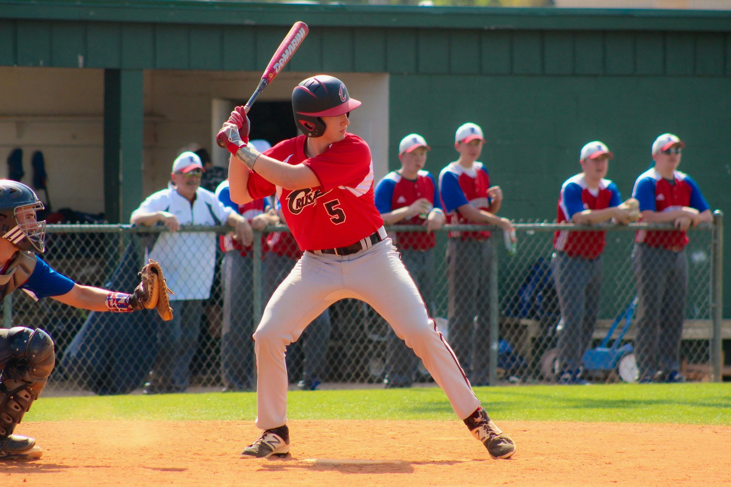 A baseball player wearing a red jersey with the number 5 at bat, with teammates and coaches watching behind a chain-link fence.