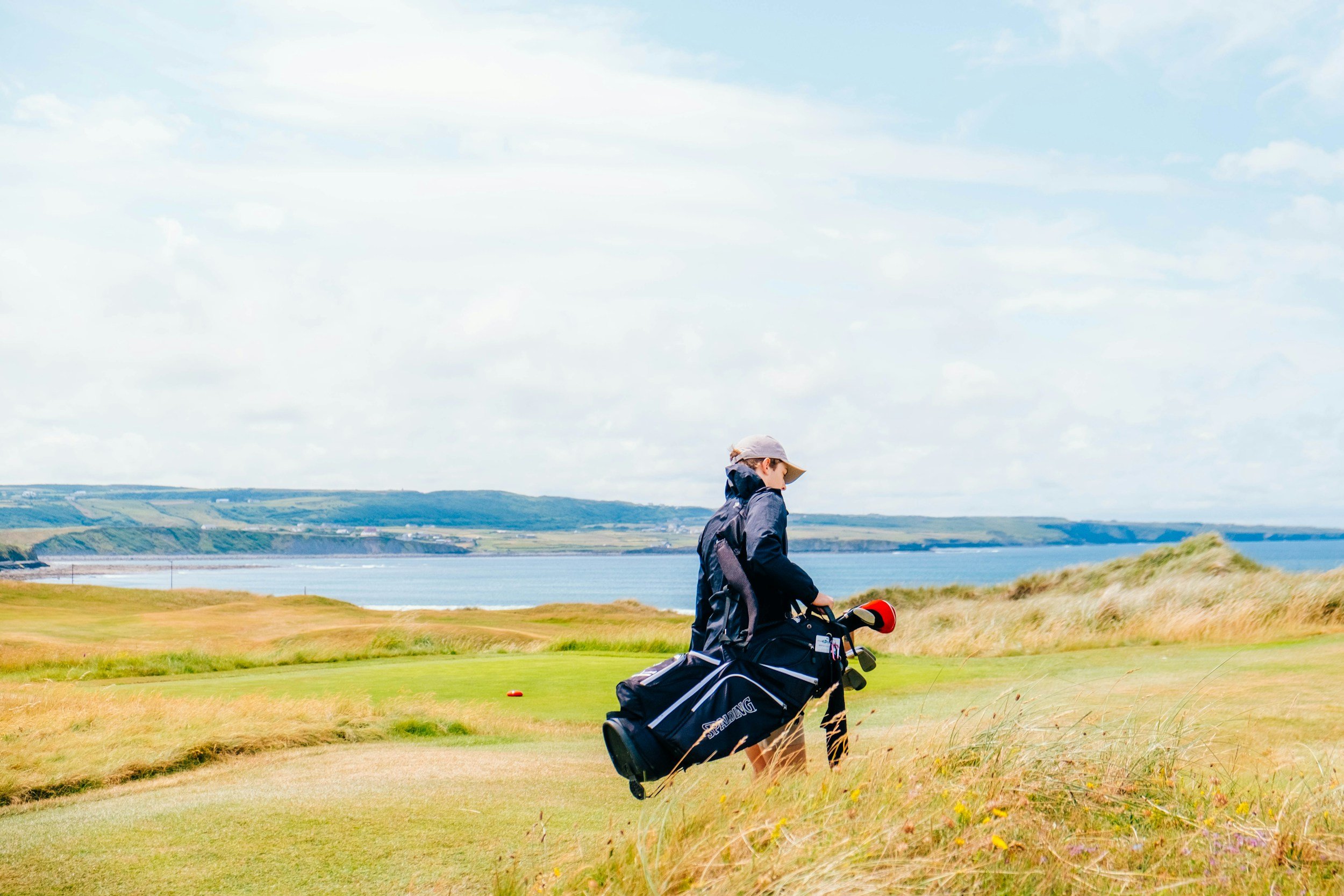 A person wearing a hat and jacket walking on a grassy golf course, carrying a golf bag, with water and hills in the background under a partly cloudy sky.