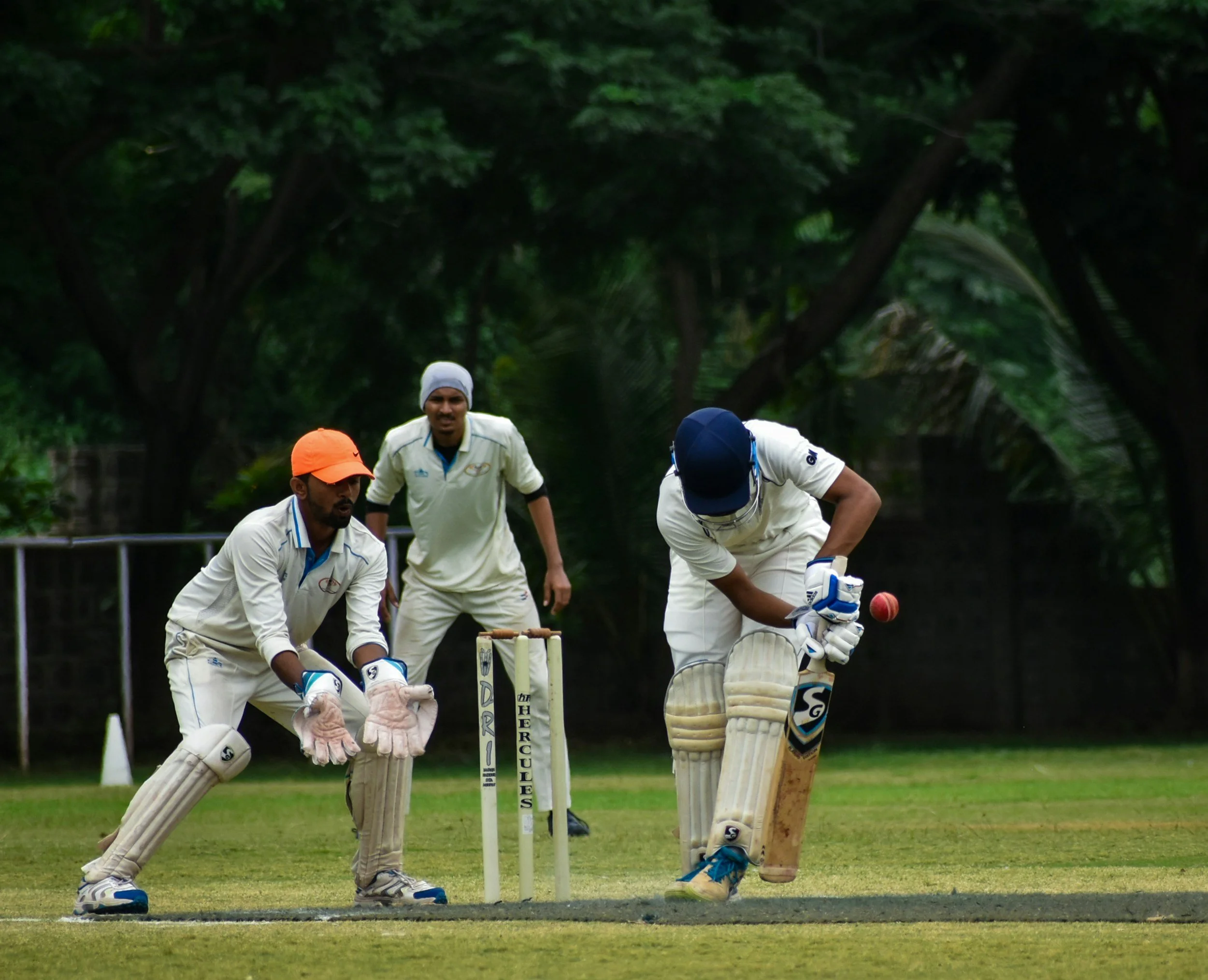 Cricket players on a field, one batsman is hitting the ball while two fielders are watching, surrounded by green trees.