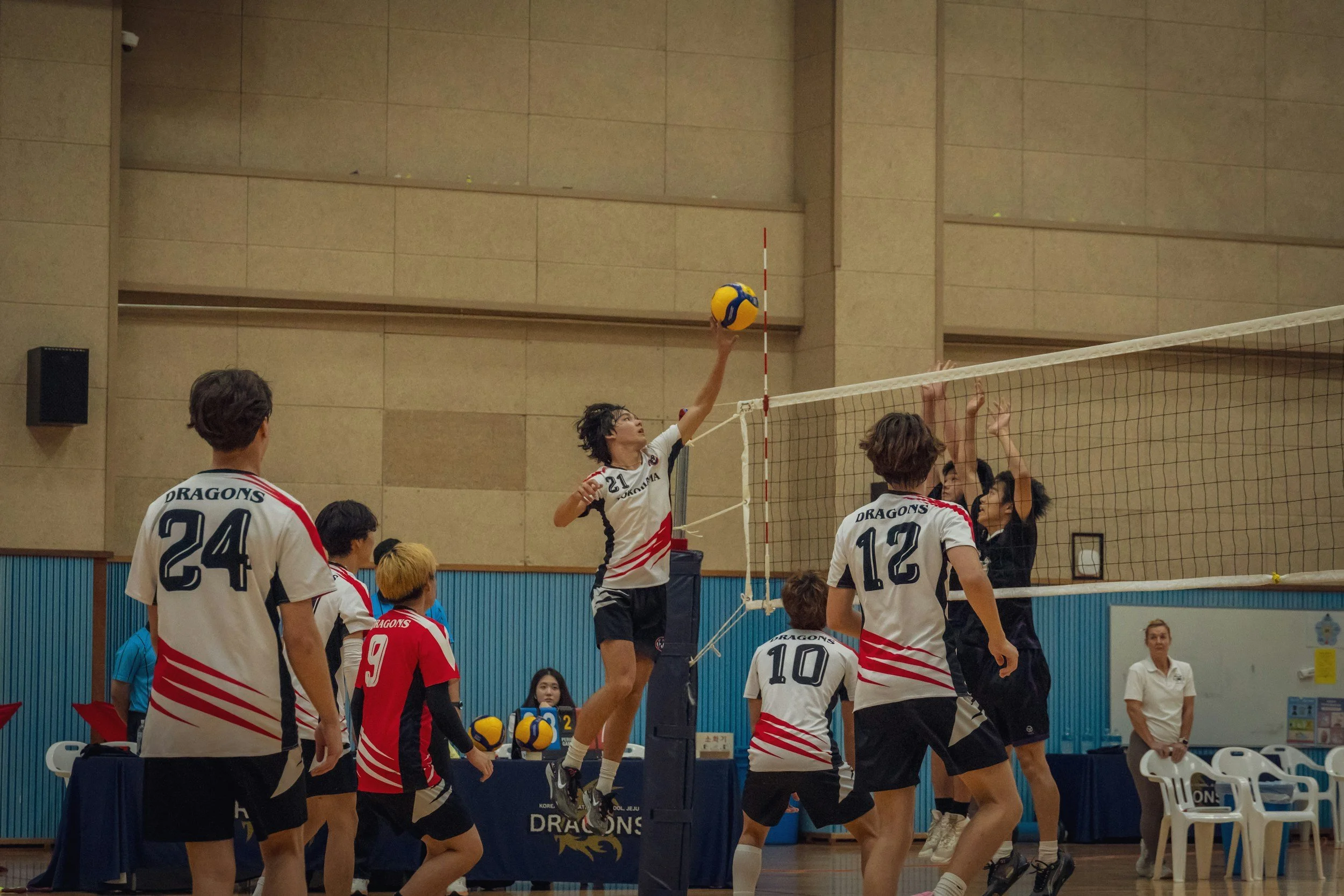 Indoor volleyball game with players jumping to hit the ball over the net, teams wearing white and red, eyes on the ball, gymnasium setting.