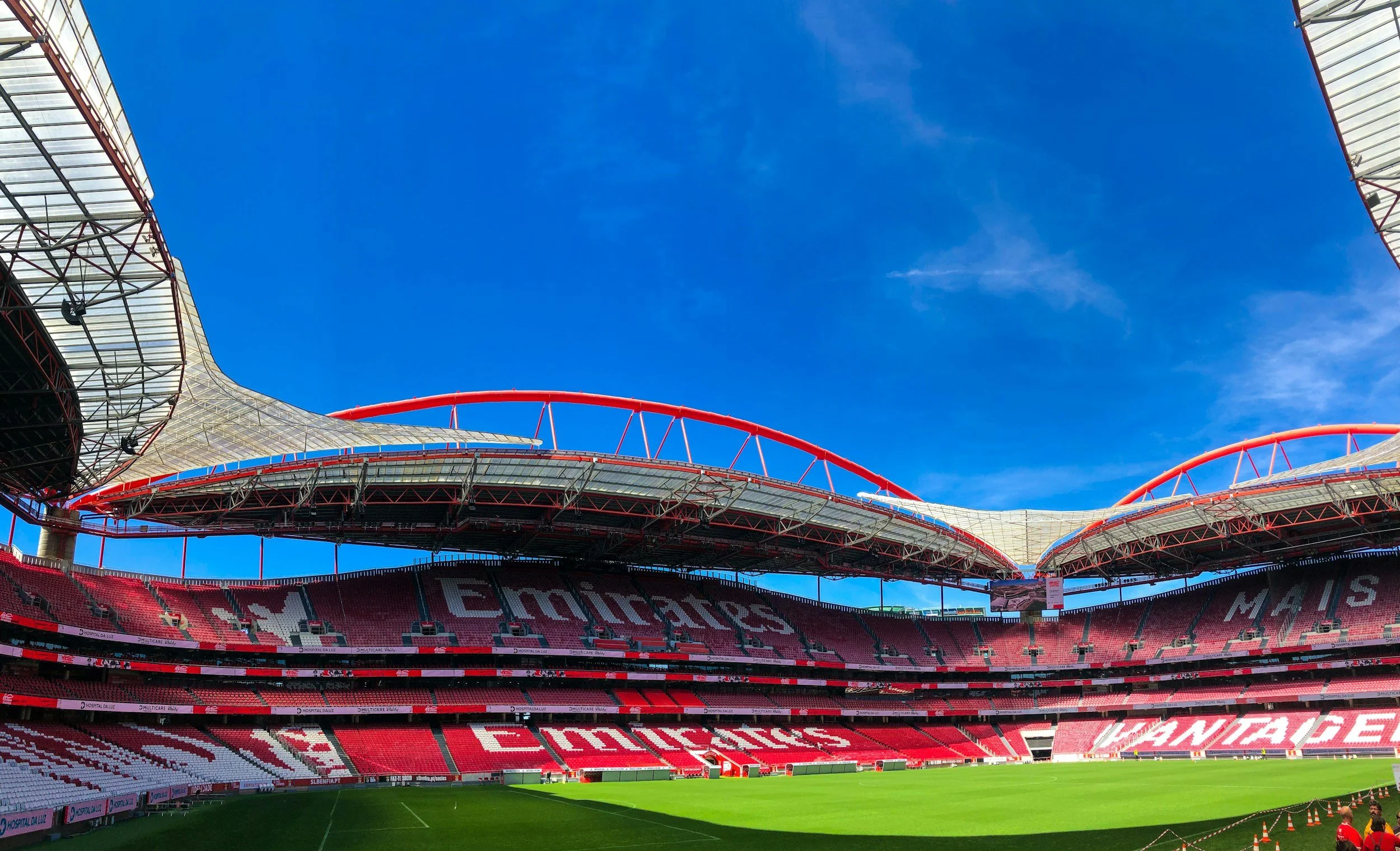 Inside view of a large football stadium with red and white seating, a green field, and a modern roof design against a blue sky.