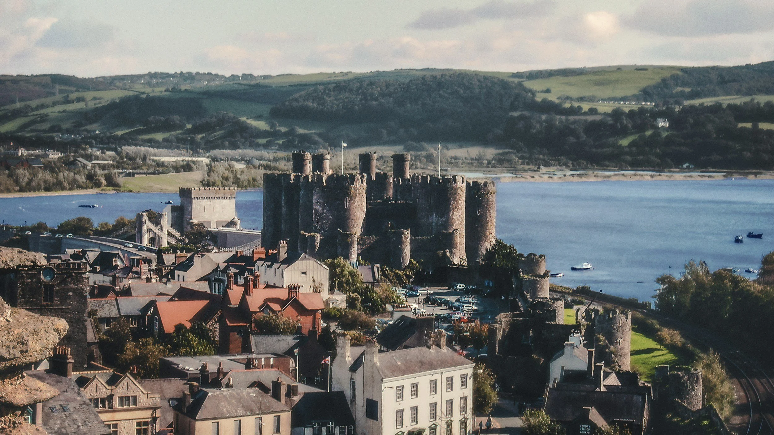 A castle by a body of water with boats, clustered houses, and a train track nearby, set against rolling green hills under a partly cloudy sky.