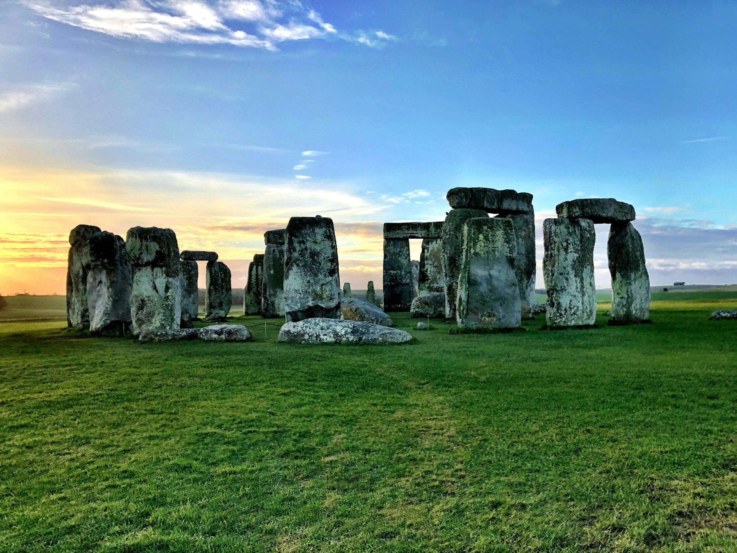 Sunset over the ancient stone monument of Stonehenge with a green grass field in the foreground and a partly cloudy sky.