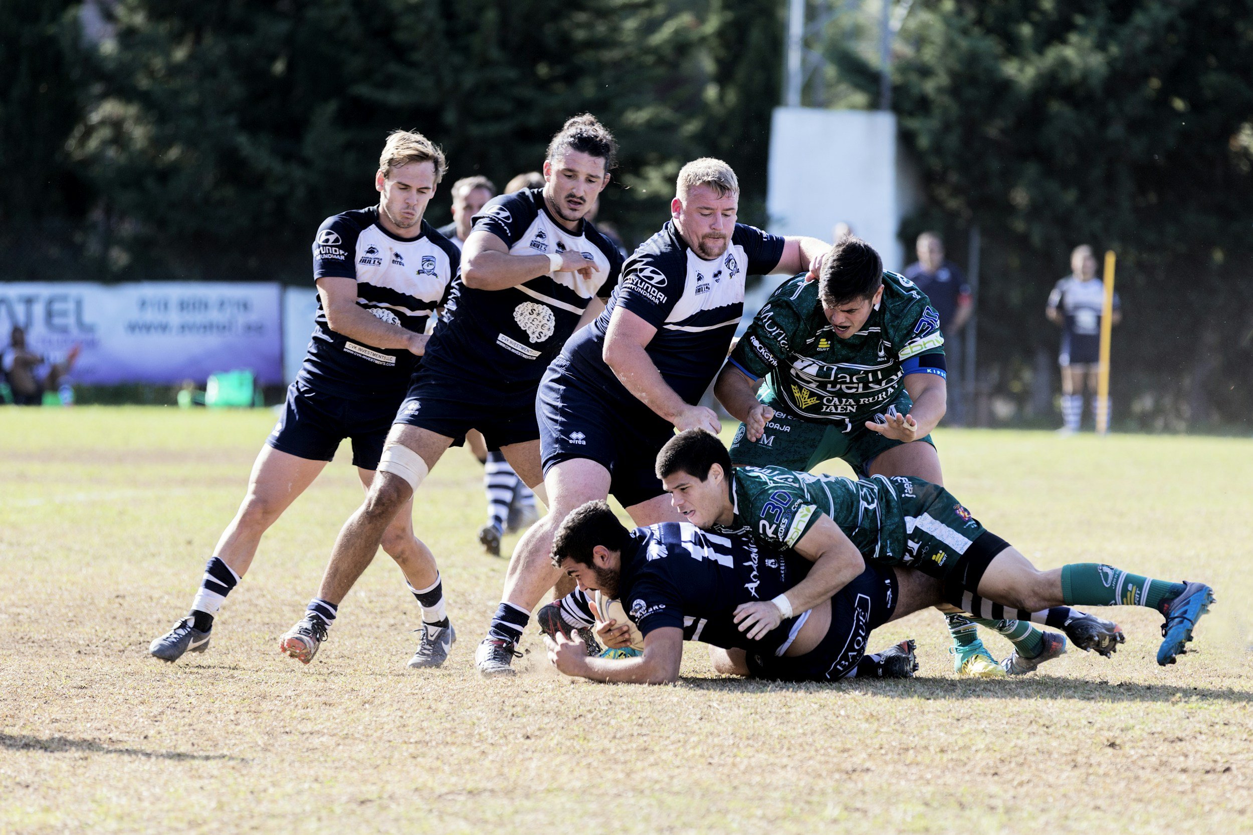 Rugby players in black and green jerseys competing for the ball during a match on a grassy field.