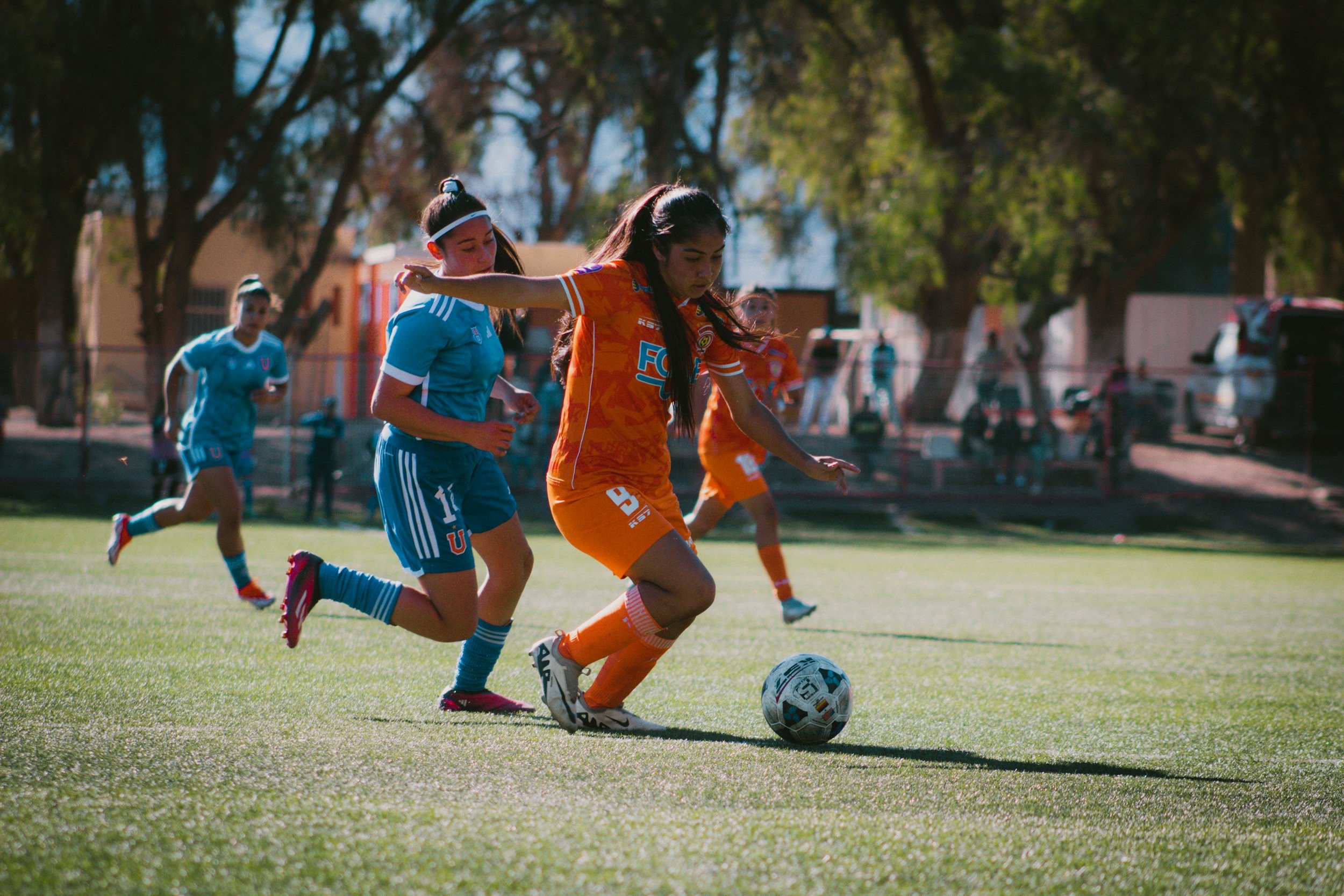 Girls playing soccer on a field, with one girl in an orange uniform kicking the ball while other girls in blue uniforms chase after her during daytime.