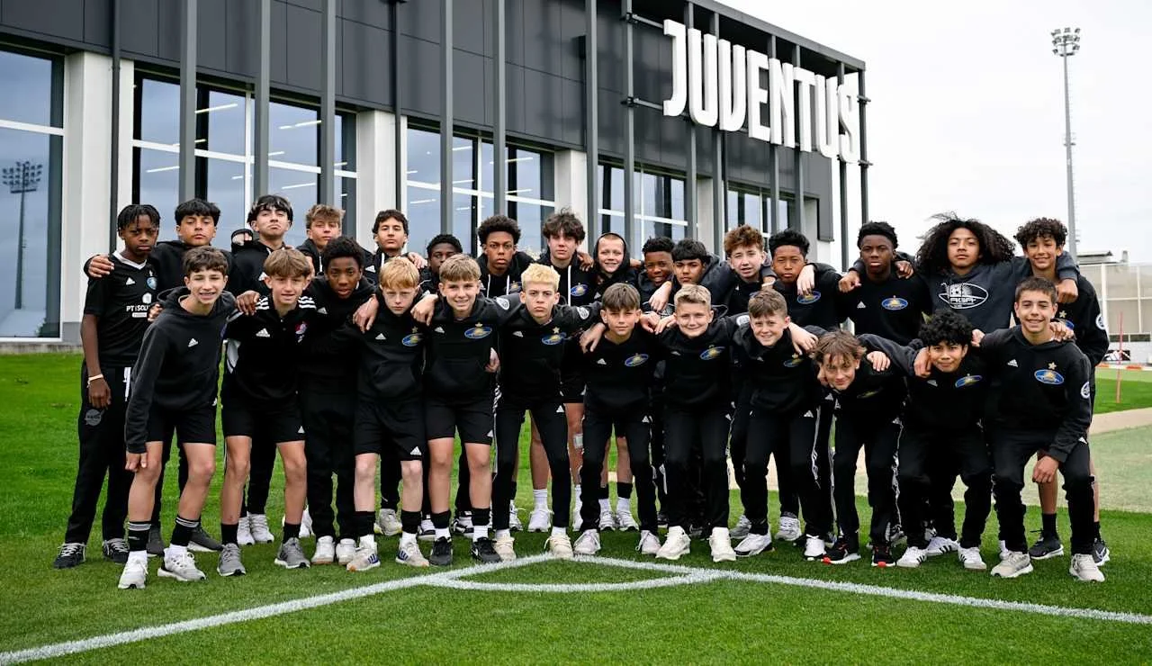 Group of young soccer players in black uniforms standing outside in front of a building with a large Juventus sign, on a green field.