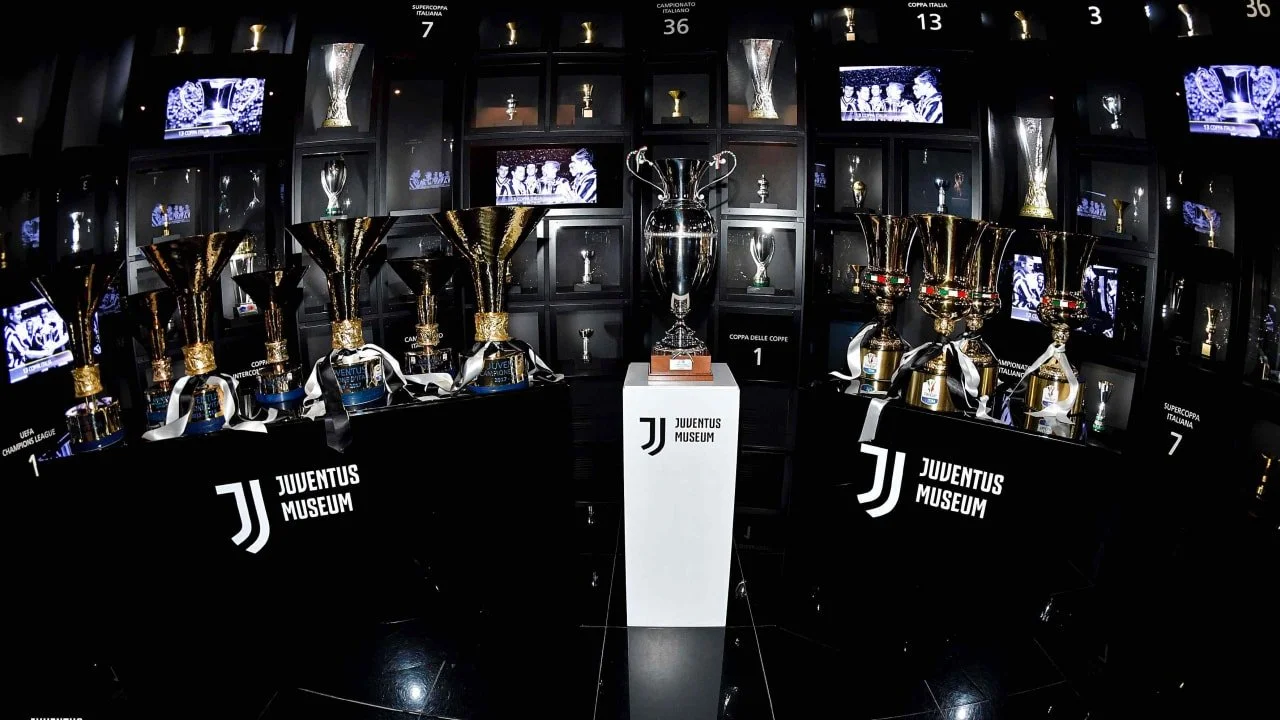 A display of multiple sports trophies, including large gold cups and a central silver trophy, at Juventus Museum with black shelving and digital screens showing game footage.