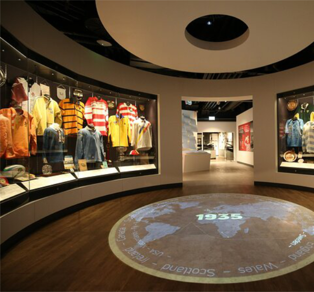 Museum exhibit featuring vintage clothing on display cases, a world map projection on the floor with the year 1935, and a modern interior with curved display walls.