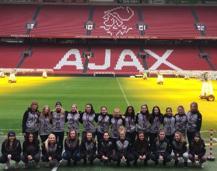 Group of female athletes in grey and black sportswear standing on a football field inside Ajax stadium.
