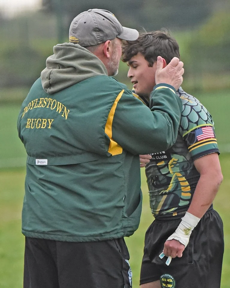 A coach and a young rugby player share an emotional moment, with the coach holding the player's face and both smiling. The coach wears a green jacket with 'POLLESTOWN RUGBY' on the back, and the player is in a camouflage rugby uniform with an America