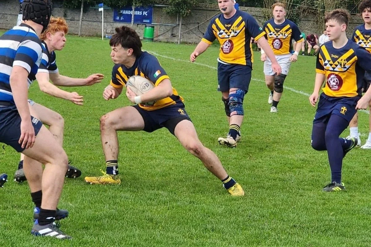 Young rugby players in yellow and blue uniforms compete during a game, with one player holding the ball and others trying to block or tackle on a grassy field.