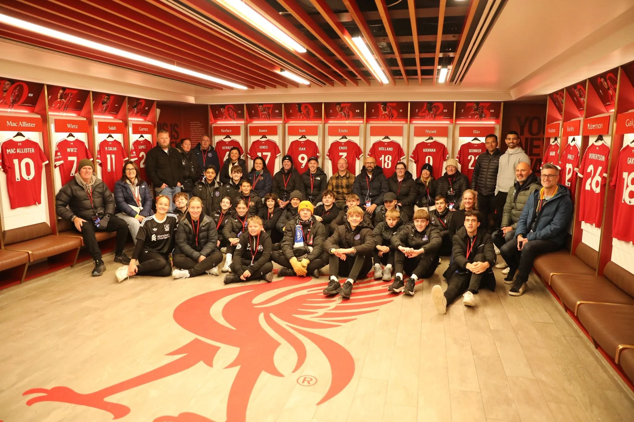 A group of children and adults posing for a photo inside a football team locker room, with red jerseys hanging on the wall behind them.