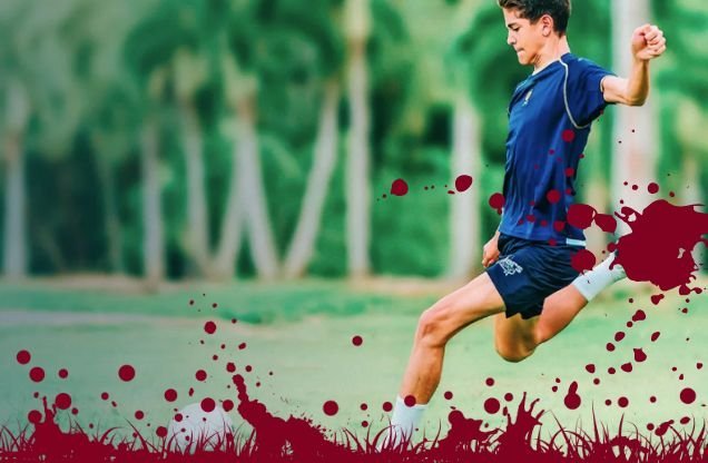 A young soccer player in a blue uniform kicks a ball on a grassy field with trees in the background, with red paint splatters over the image.