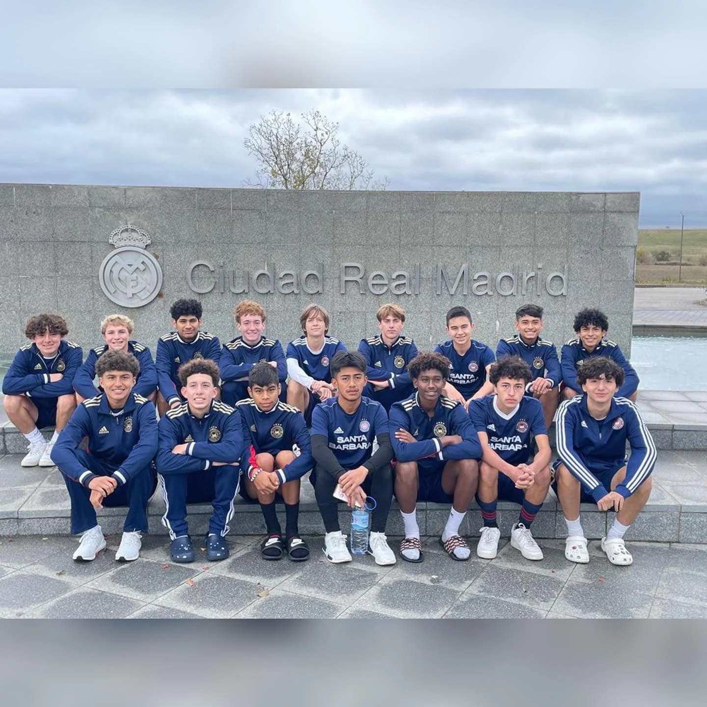 Group of young male soccer players in blue uniforms posing outdoors in front of a stone wall with the inscription 'Ciudad Real Madrid' and the Madrid football club logo.
