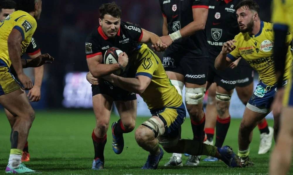 Rugby players in action during a game, with one player in black holding the ball and running past opponents in yellow and blue jerseys, on a field.
