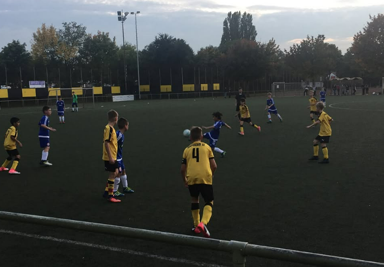 Children playing soccer on a field during daytime, some in yellow jerseys and others in blue, with trees in the background and cloudy sky.