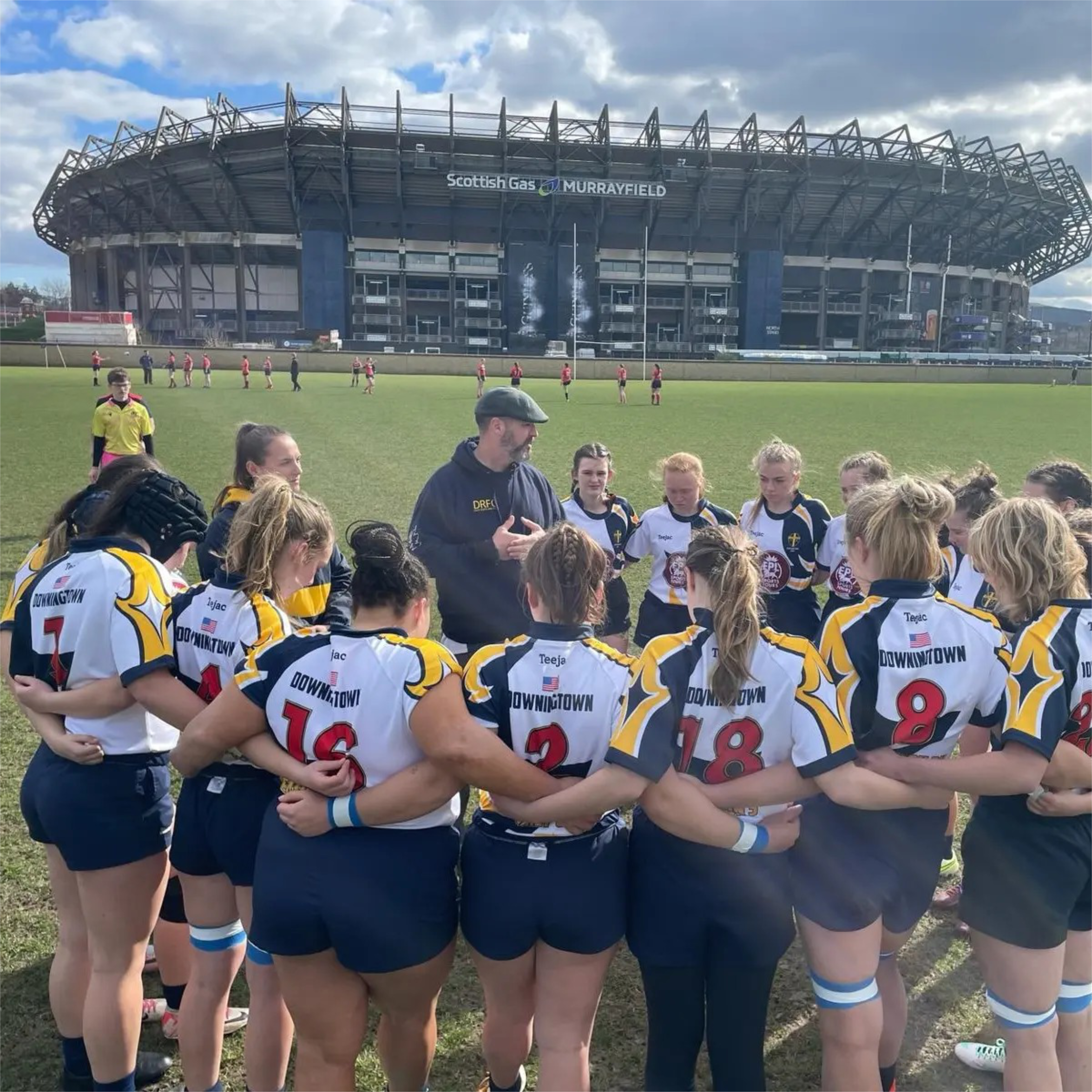 A women's rugby team huddles around their coach on a rugby field with Murrayfield Stadium in the background.