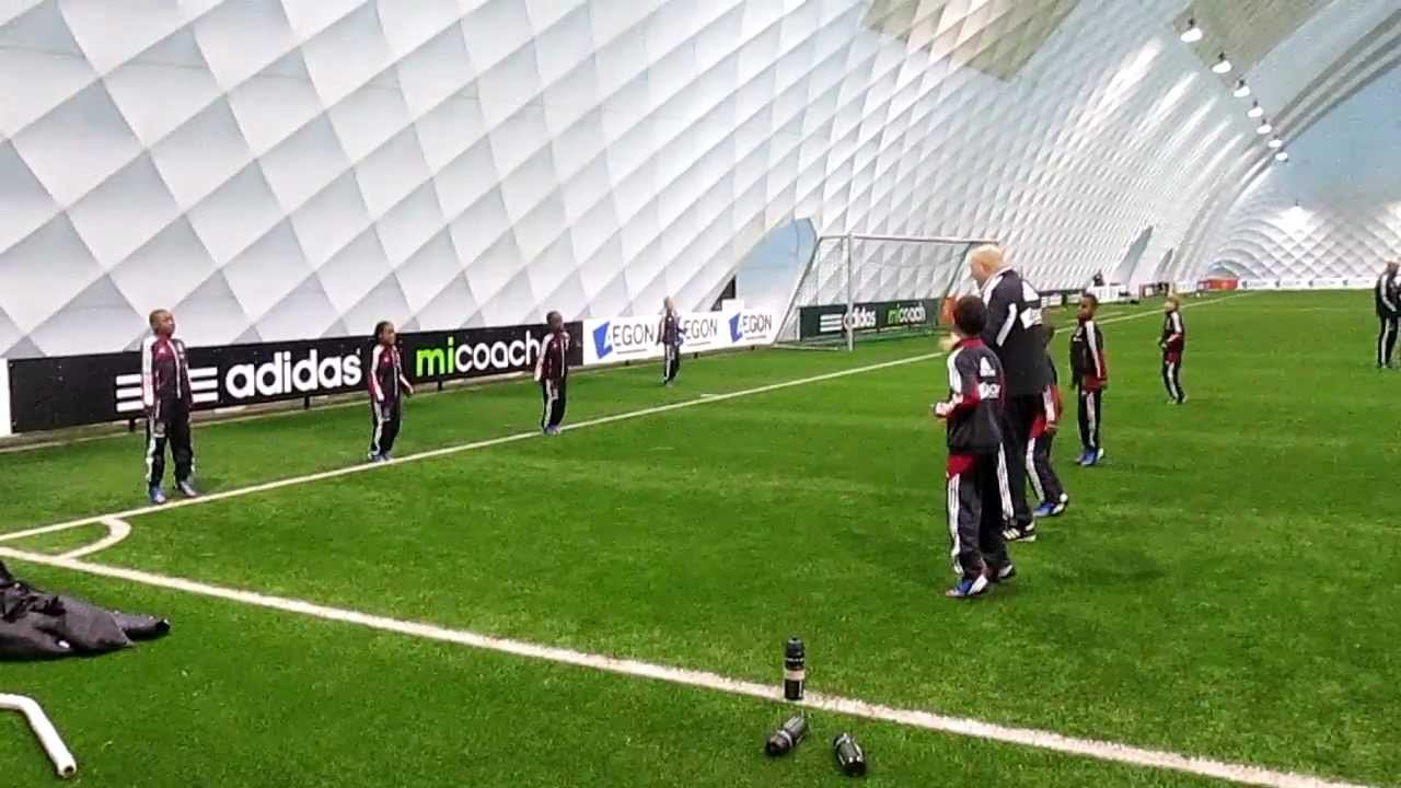 Children and coaches practicing soccer on an indoor field with artificial grass, curved interior walls, and advertising banners along the perimeter.