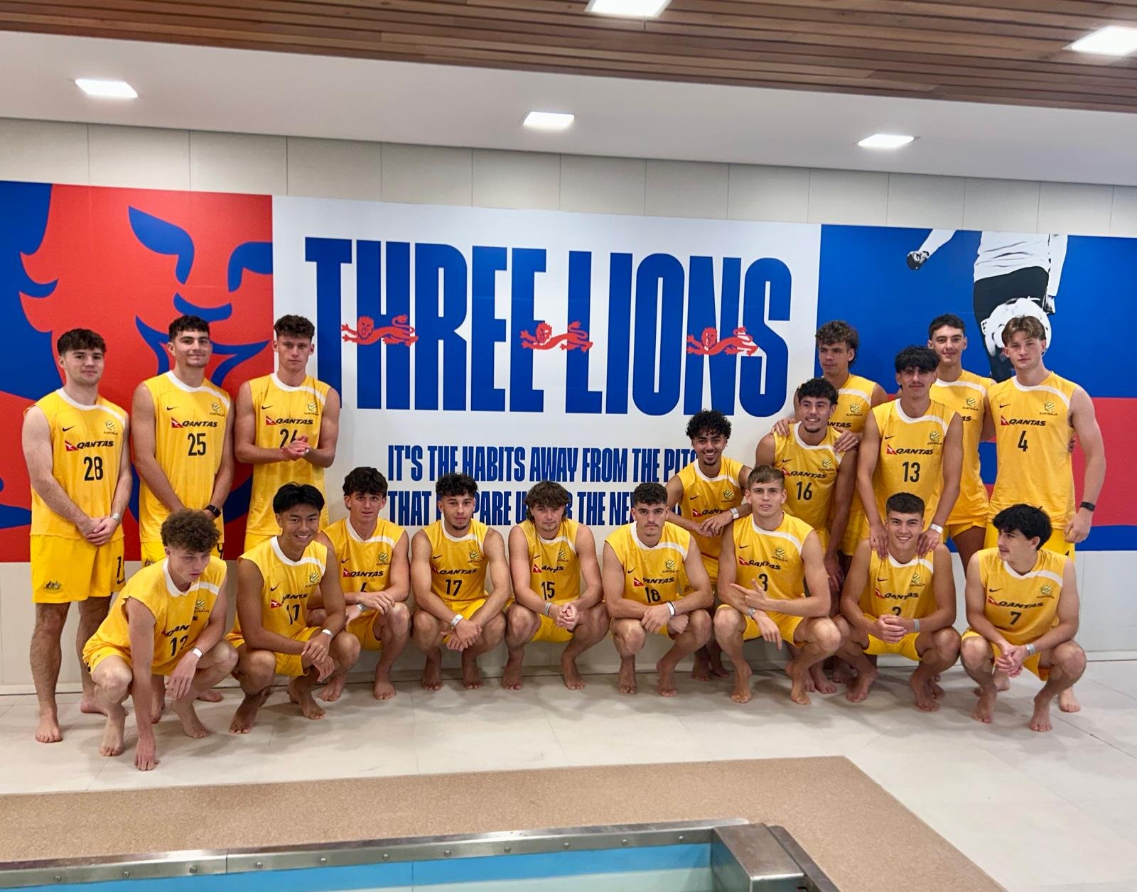 A group of young male athletes in yellow sports uniforms pose for a team photo inside an indoor pool area. They are standing and squatting in front of a large sign that reads 'TIRELLIONS' with a lion logo and a quote about habits.