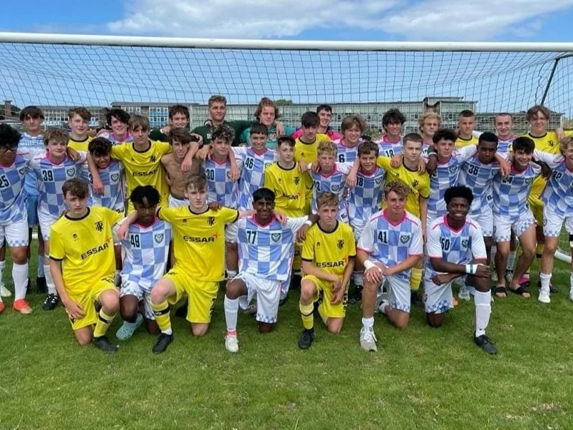 Group of young soccer players posing in front of a goal on a grassy field, some wearing yellow jerseys and others in blue and white uniforms.