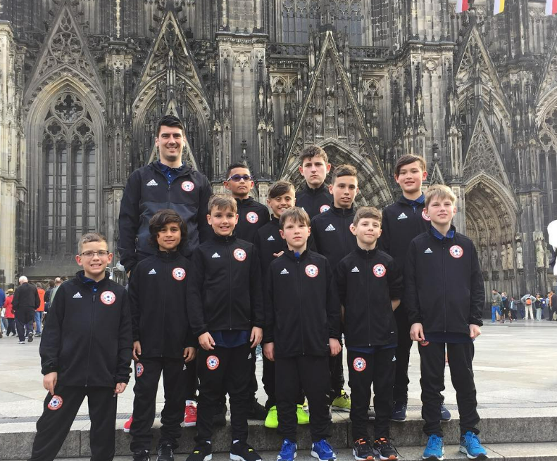 A group of young boys and their coach or chaperone standing in front of a Gothic cathedral, possibly during a sports or school trip.