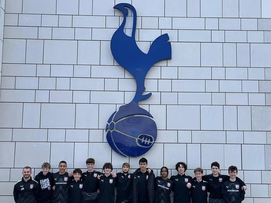 A group of young men in black soccer jackets standing in front of a large wall display of the Tottenham Hotspur football club logo, which features a blue cockerel standing on a football.