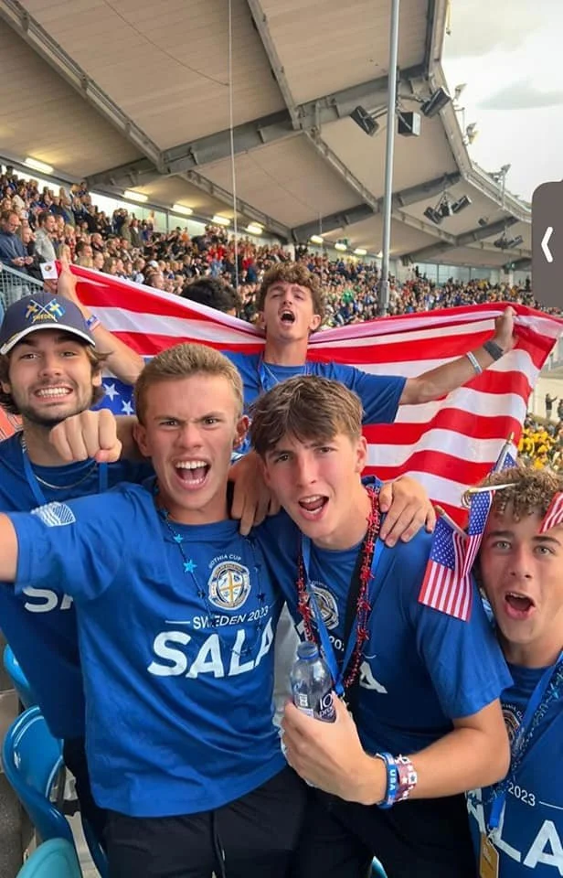 Group of young men celebrating at a sports event, holding an American flag, wearing blue shirts with a logo, and enjoying the crowd and stadium atmosphere.