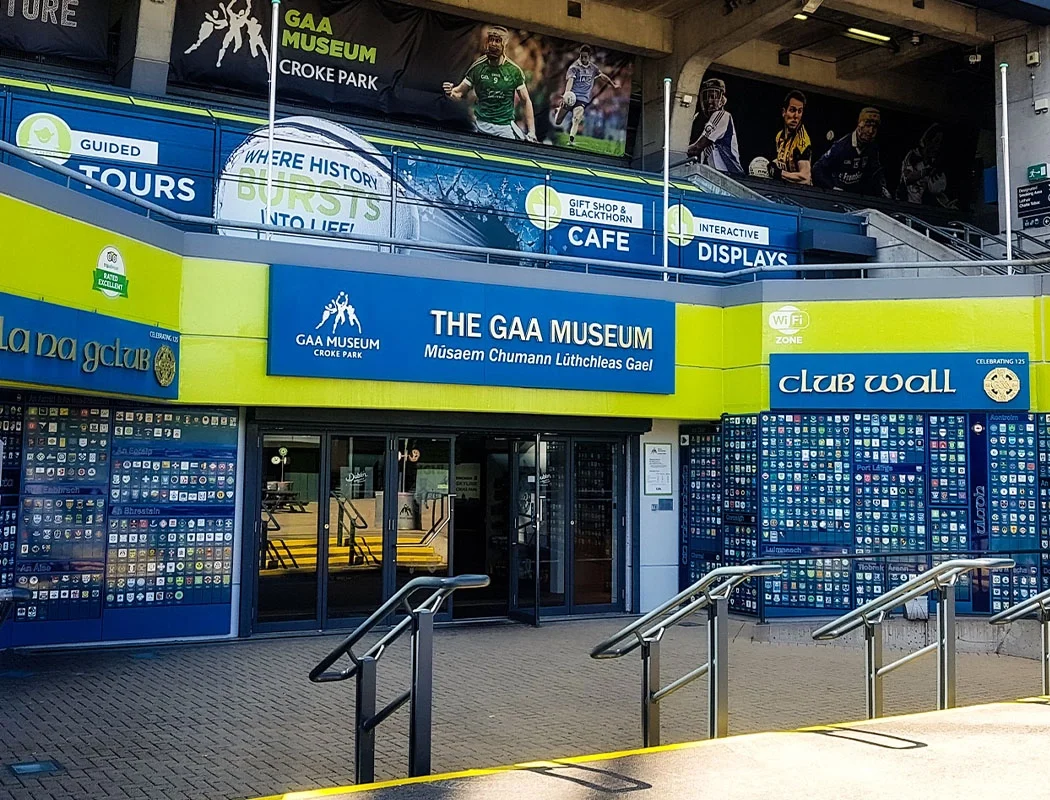 The entrance of the GAA Museum at Croke Park with blue signage and club wall, and advertisements for guided tours, the gift shop, cafe, and interactive displays.