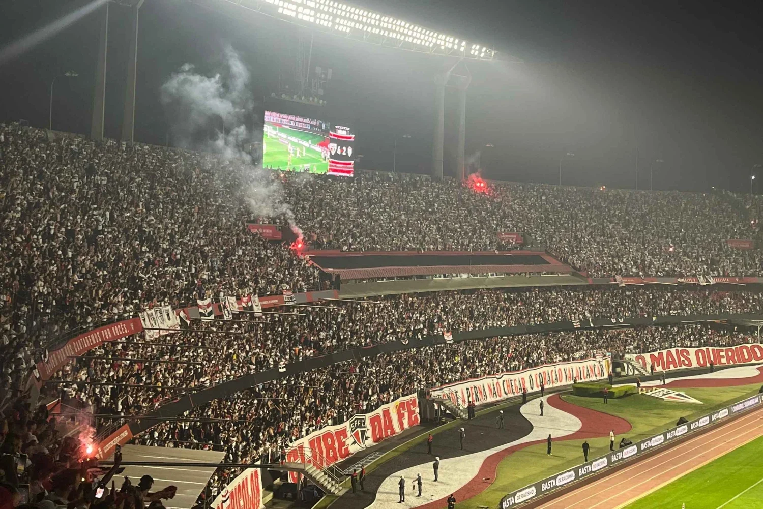 Crowd of football fans in stadium stands, some holding flags and banners, with a large video screen displaying the game, and smoke/flairs releasing red smoke among the spectators.