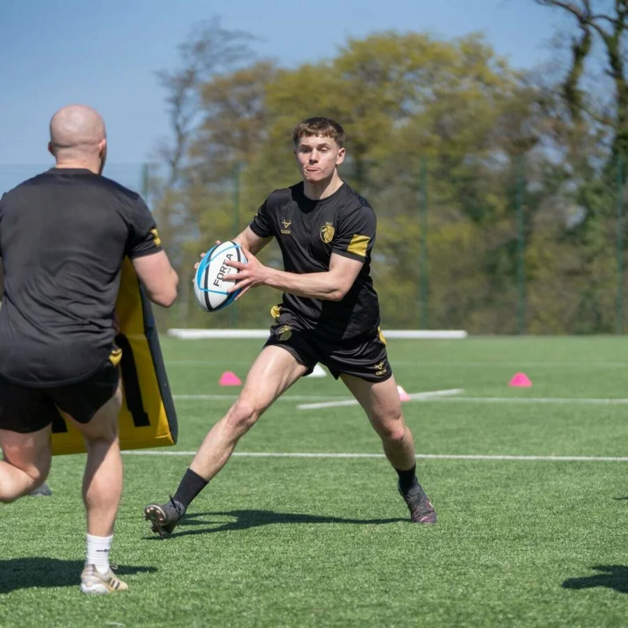 A young male rugby player in black uniform holding a rugby ball during a practice or training session on a grass field.