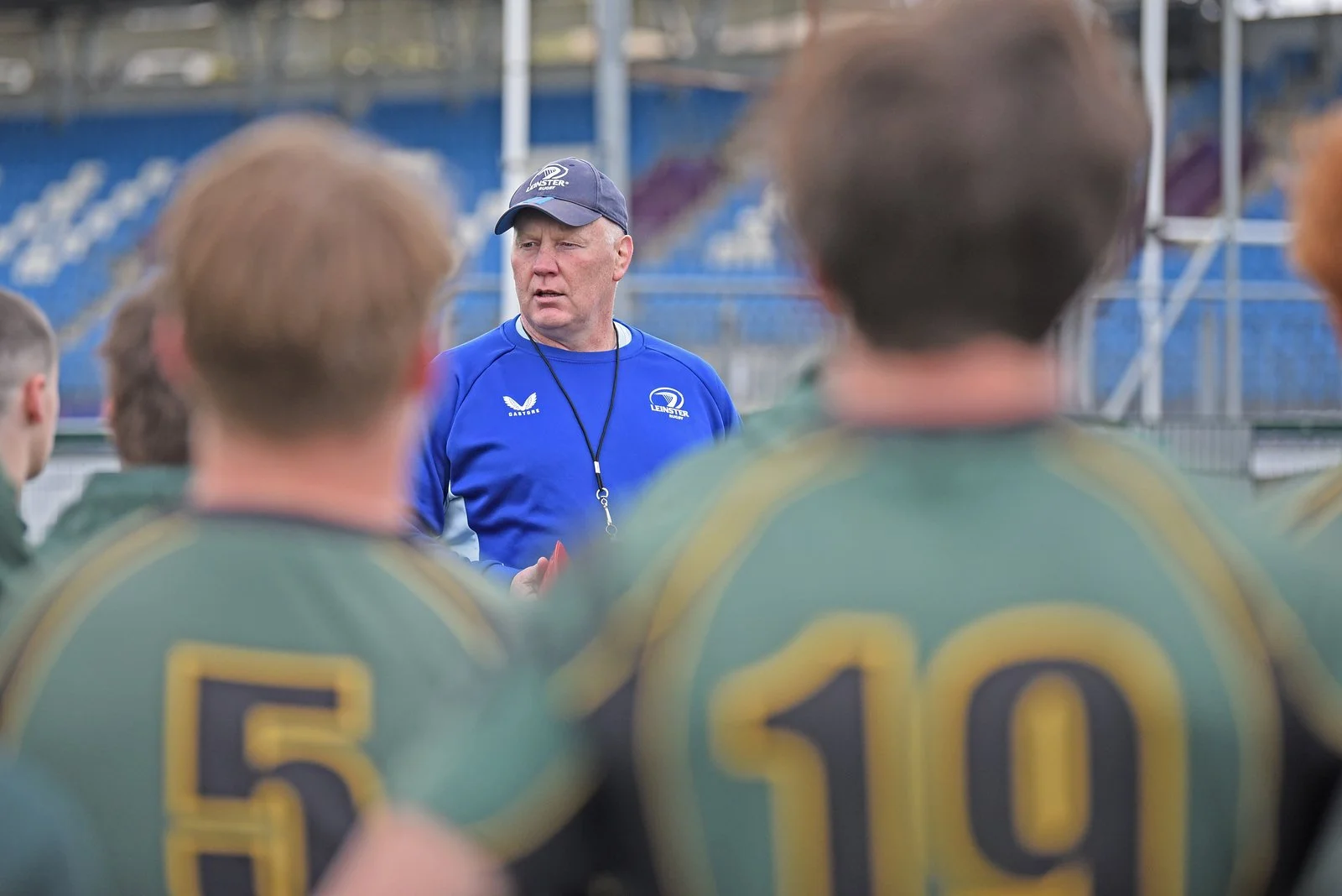A coach or referee in a blue shirt and cap speaking to a team of football players wearing green and yellow jerseys, with numbers 5 and 10 visible, on a football field.