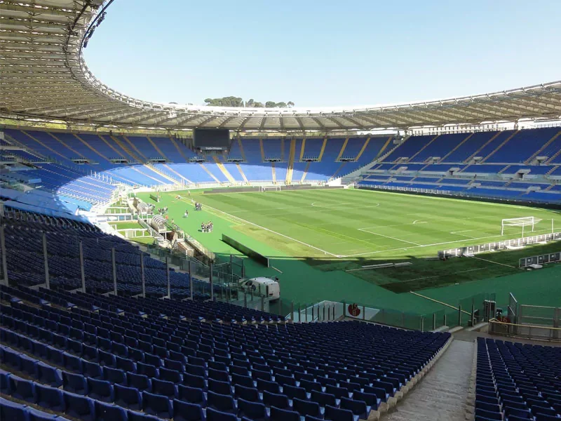 Empty football stadium with blue seats and a green field under a clear sky.