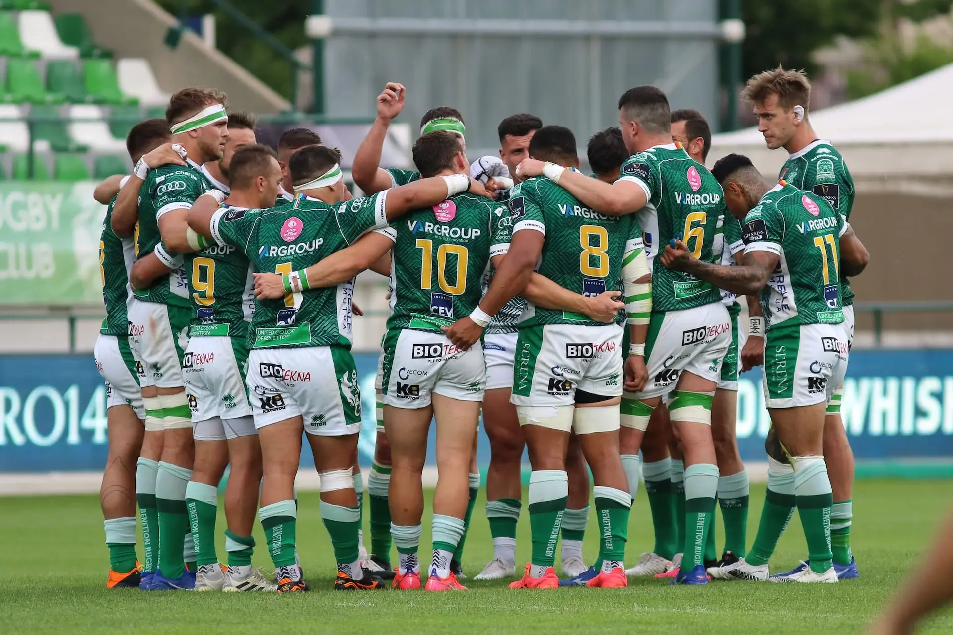 A rugby team in green and white uniforms huddles together on the field during a match.