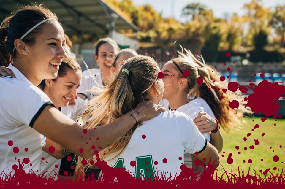 Group of young female soccer players celebrating and hugging each other on a sunny outdoor field, with trees and bleachers in the background.