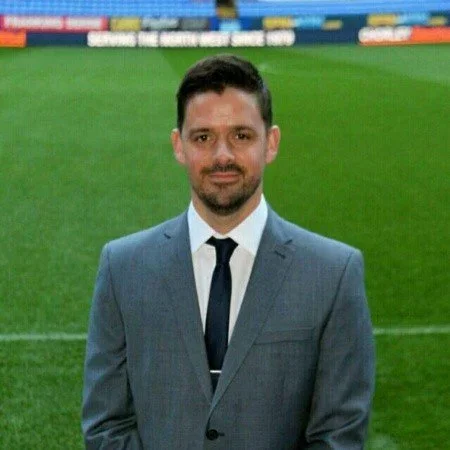 A man in a gray suit and tie standing on a sports field with a green grass background.