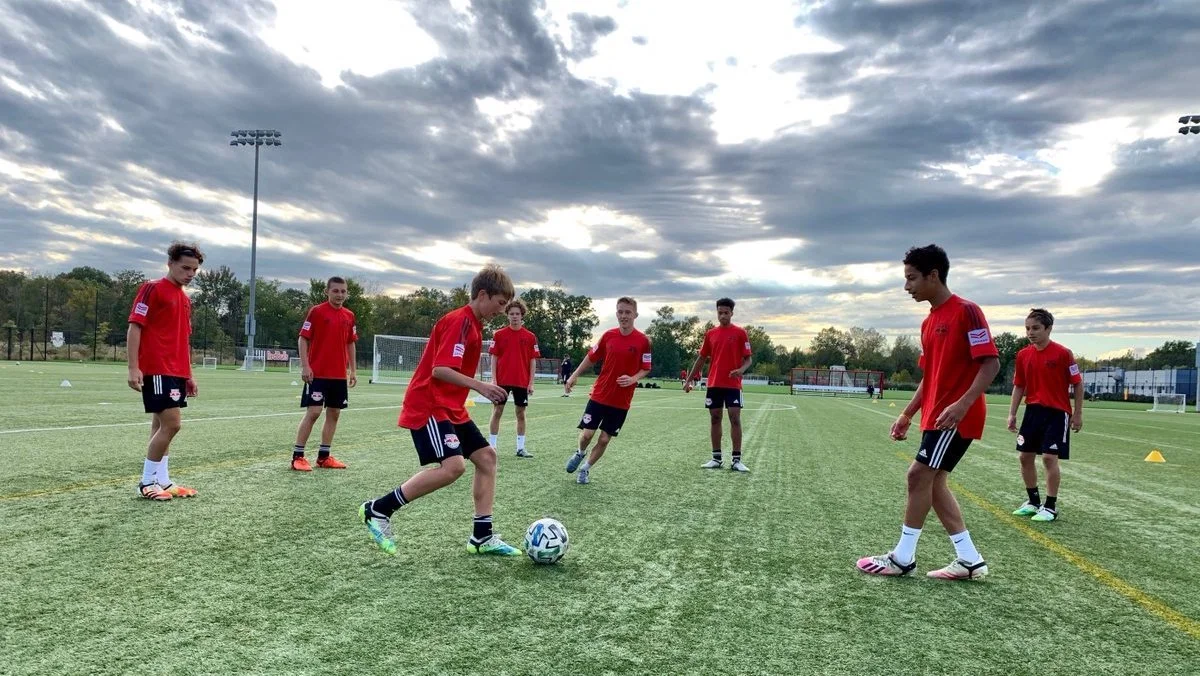 A group of young boys practicing soccer on a field under a cloudy sky, with some wearing red jerseys and others in black shorts.