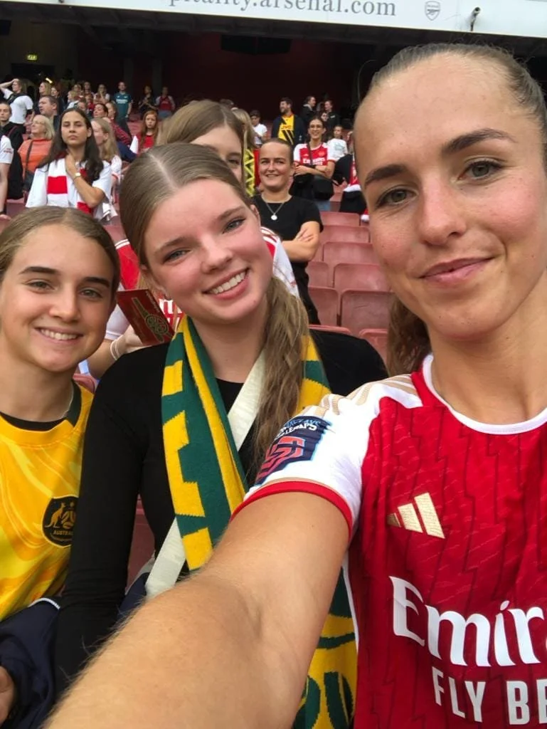 Group of women at a sports event, some wearing soccer jerseys and scarves, smiling for a selfie with stadium seats and more spectators in the background.