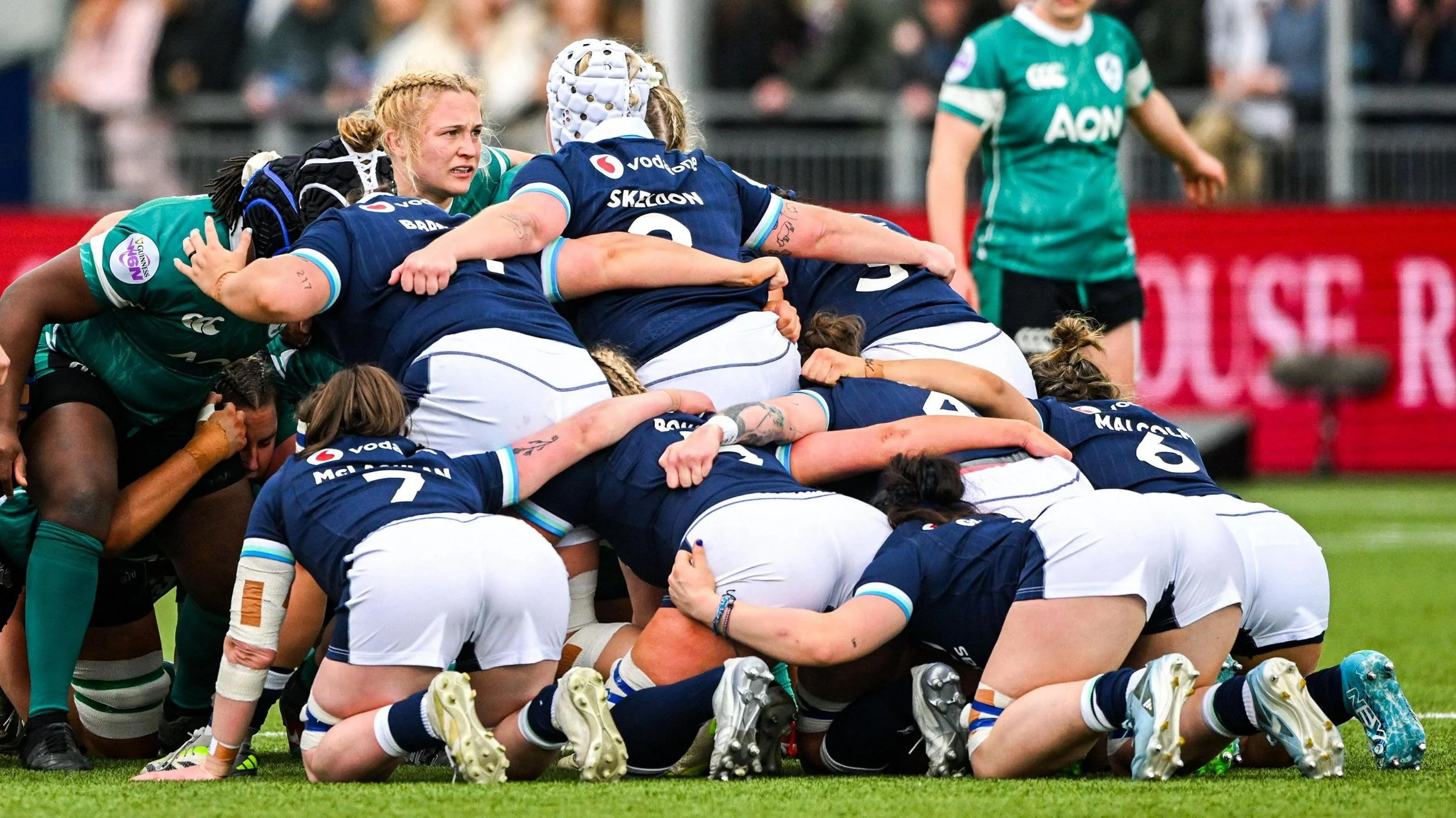 A group of female rugby players in a scrum on a rugby field, with some players wearing navy blue jerseys and others in teal jerseys, during a game.