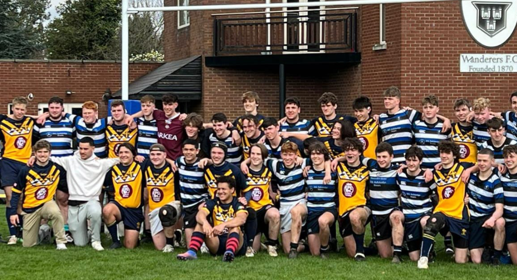 A group of young male rugby players with a coach, posing for a team photo outside a brick building with a sign that says 'Wanderers F.C., Founded 1870'.