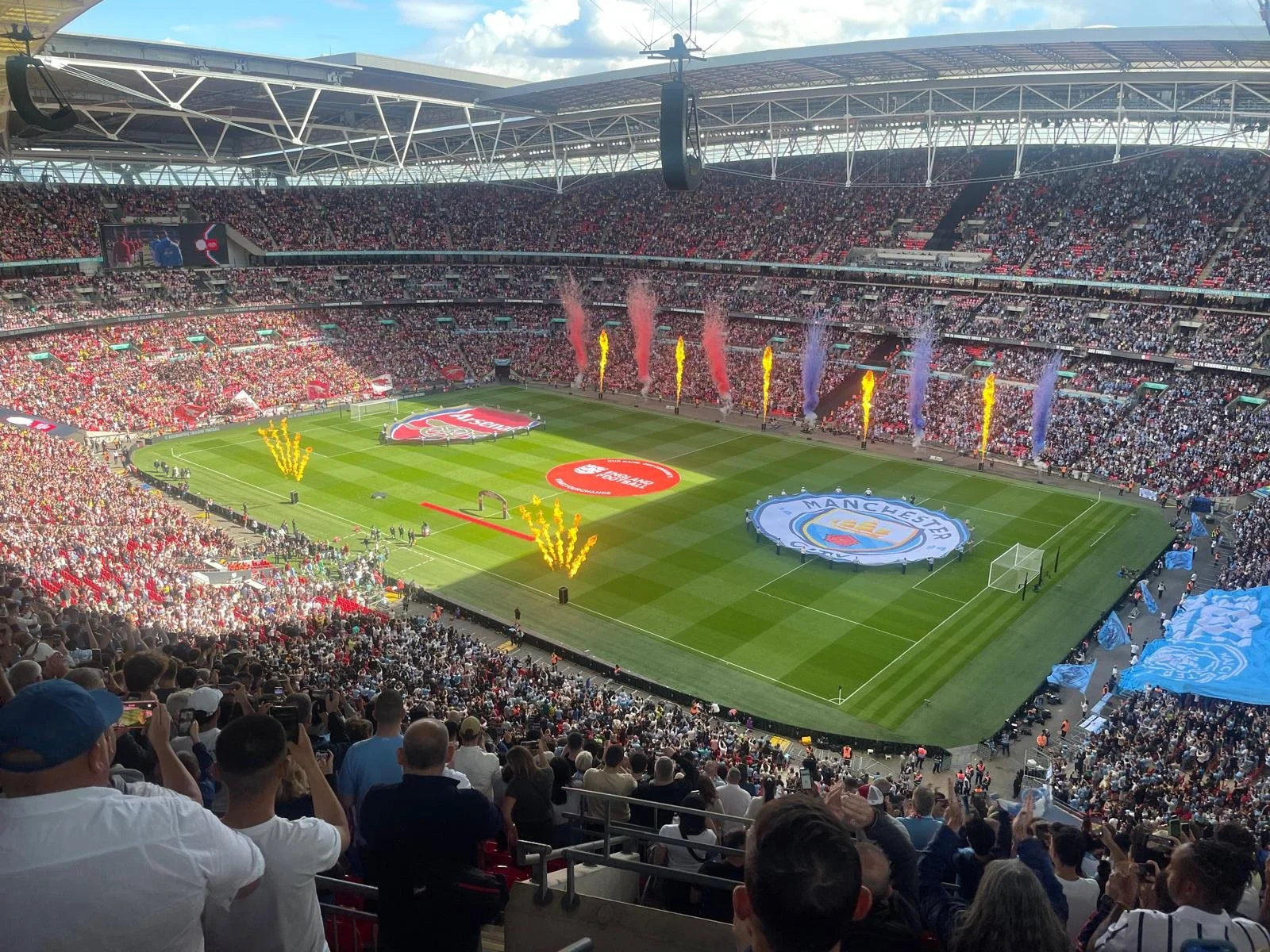 A packed soccer stadium with fans, banners, and fireworks, celebrating before a match between Arsenal and Manchester City.