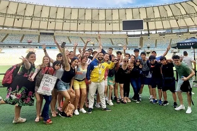 Group of young people posing and smiling on a sports field, some holding signs and making peace signs. The setting is a stadium with empty stands and a large screen overhead.