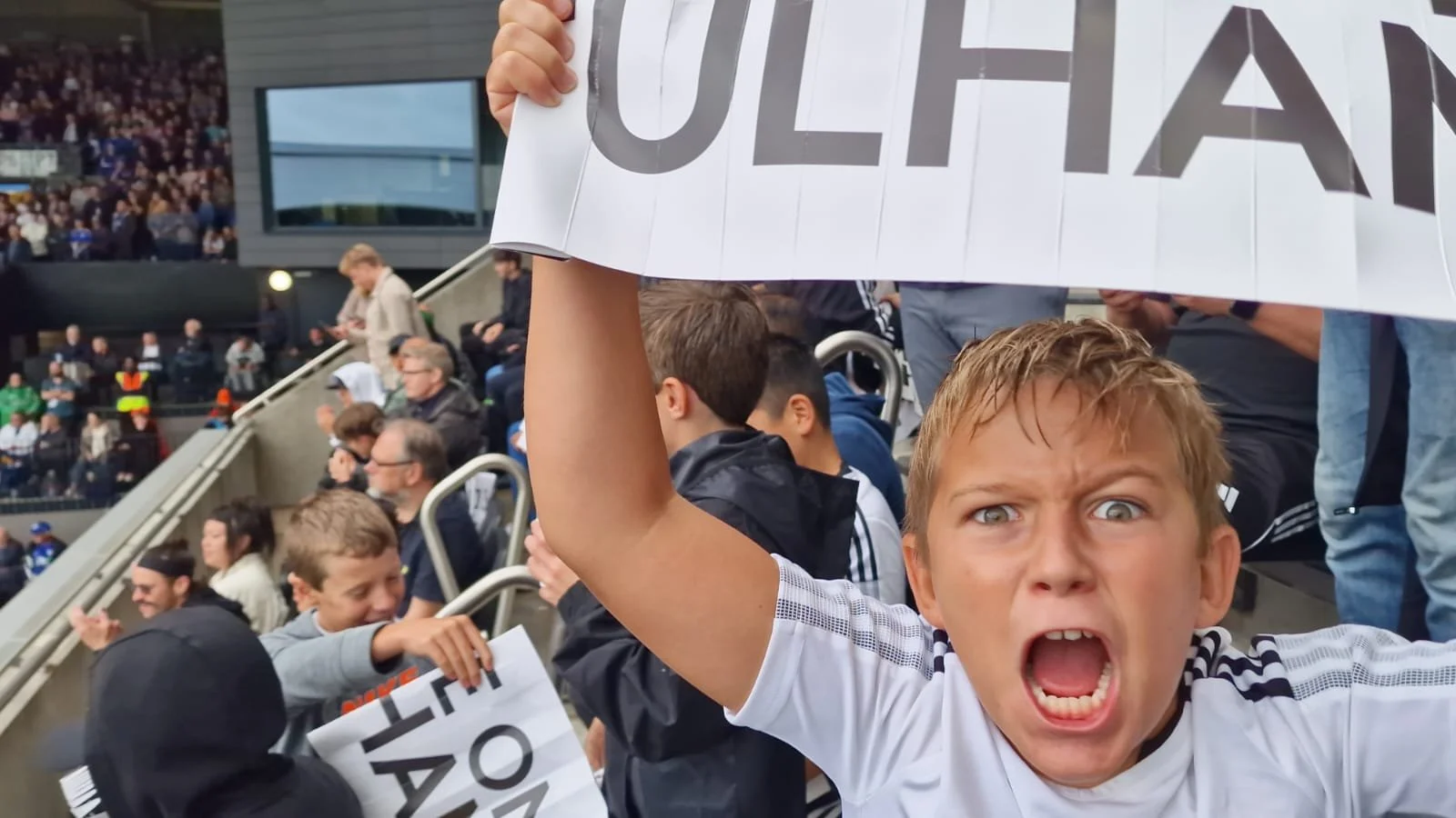 A young boy with an angry expression, mouth open as if shouting, holding a sign with large partially visible text at a sports event with many spectators in the background.