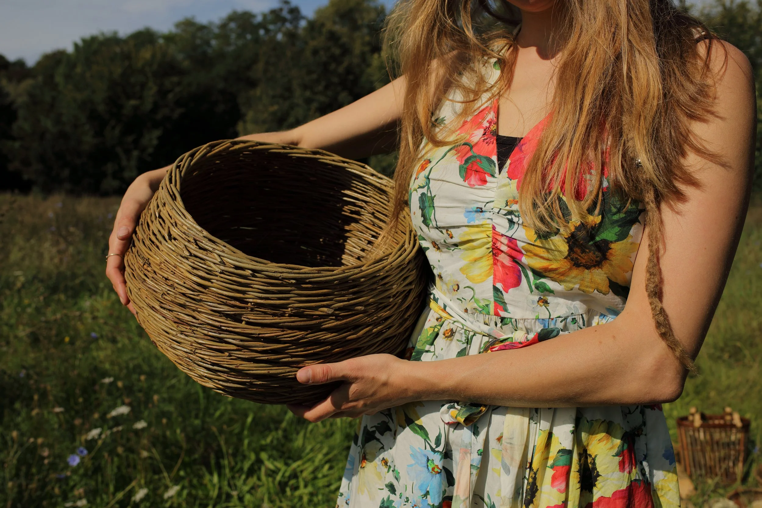 Mujer con vestido floral sosteniendo una cesta de mimbre en un campo al aire libre.