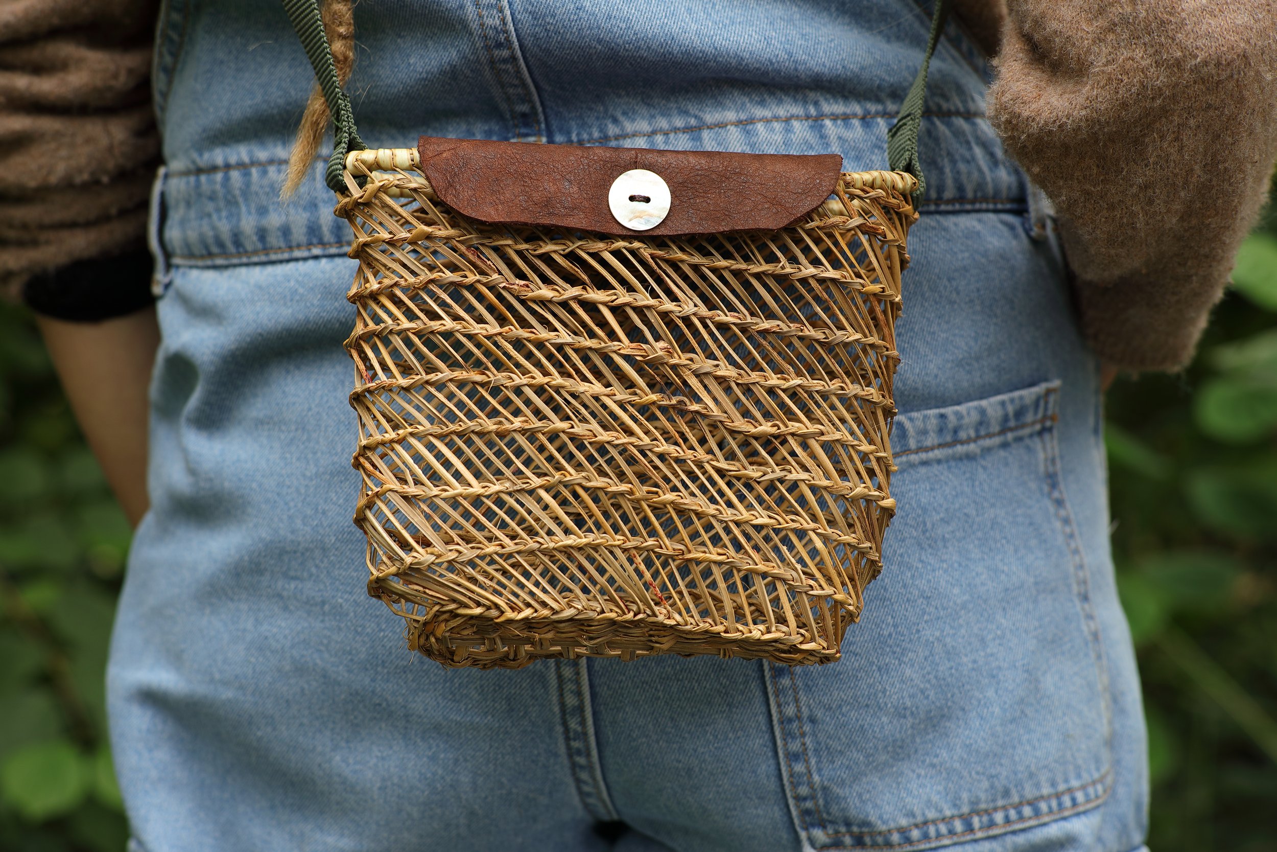 Persona con pantalones vaqueros y suéter, llevando una bolsa de mimbre colgada alrededor de la cintura, en un entorno natural.