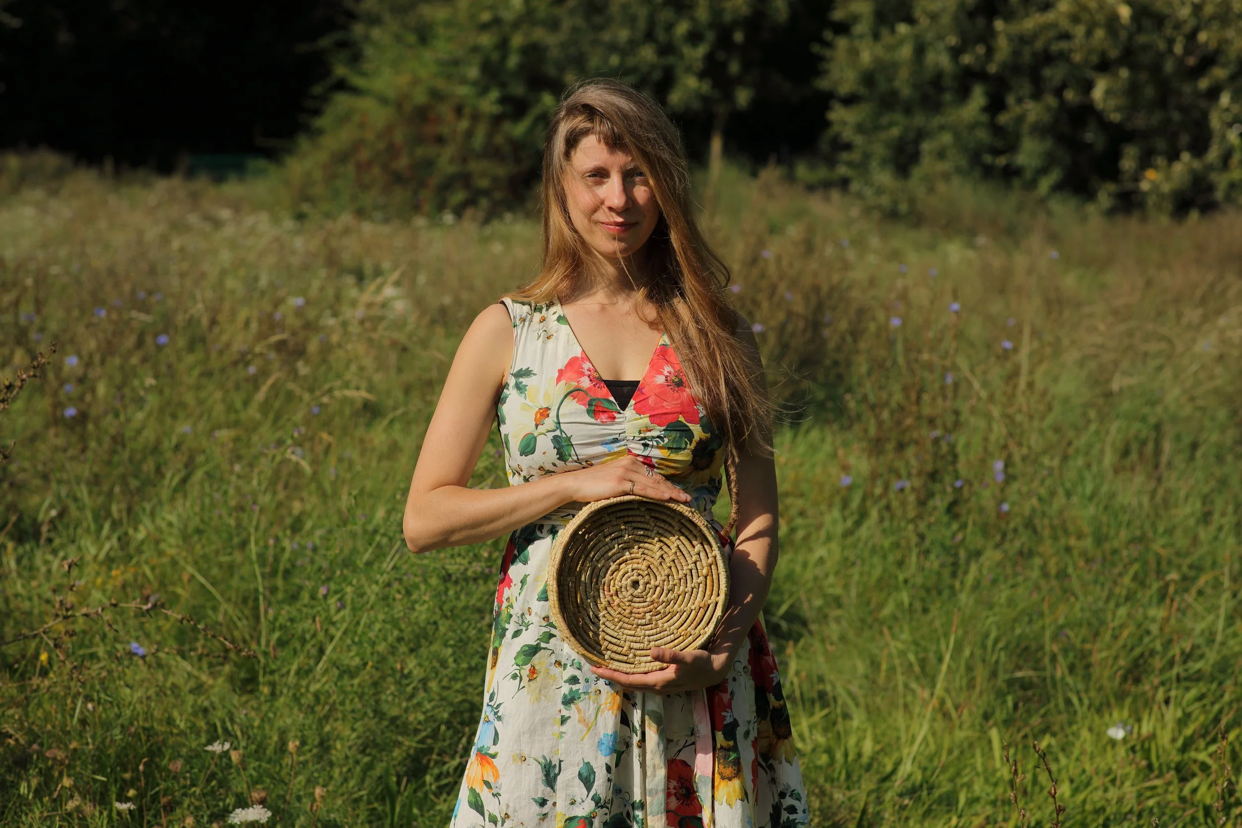 Mujer en un campo de hierba con vestido floral, sosteniendo una cesta de mimbre en un entorno natural al aire libre.