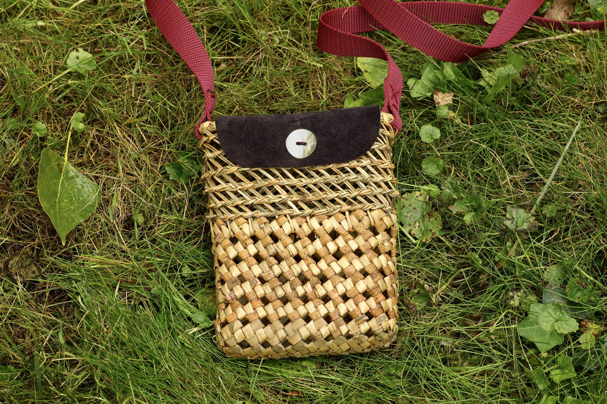 Bolso de pita phormium tenax con correa de tela y botón, colocado sobre césped verde con algunas hojas.