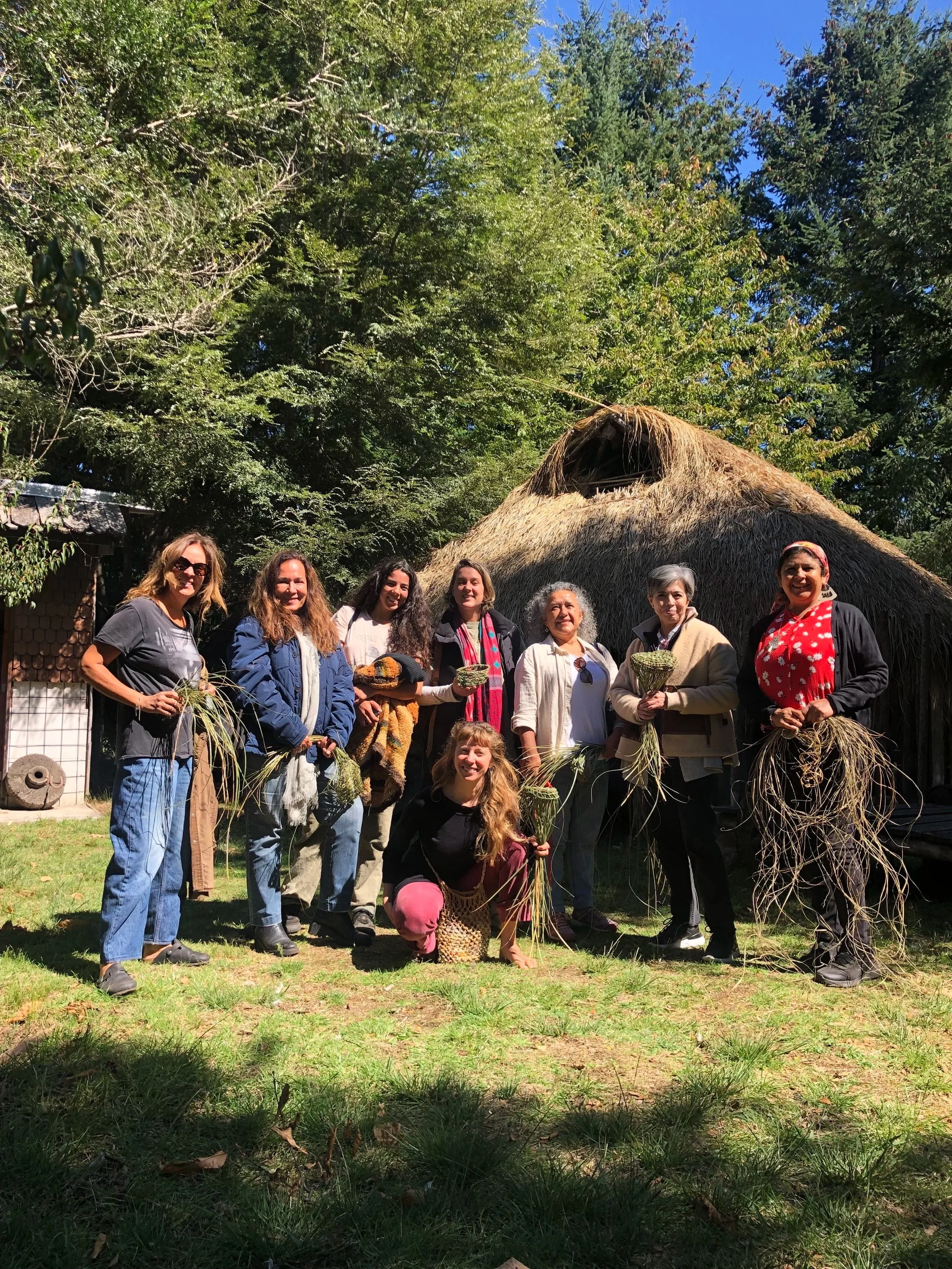 Grupo de personas en un entorno rural con un techo de paja, tomando parte en una actividad agrícola o cultural, sosteniendo plantas o hierbas.
