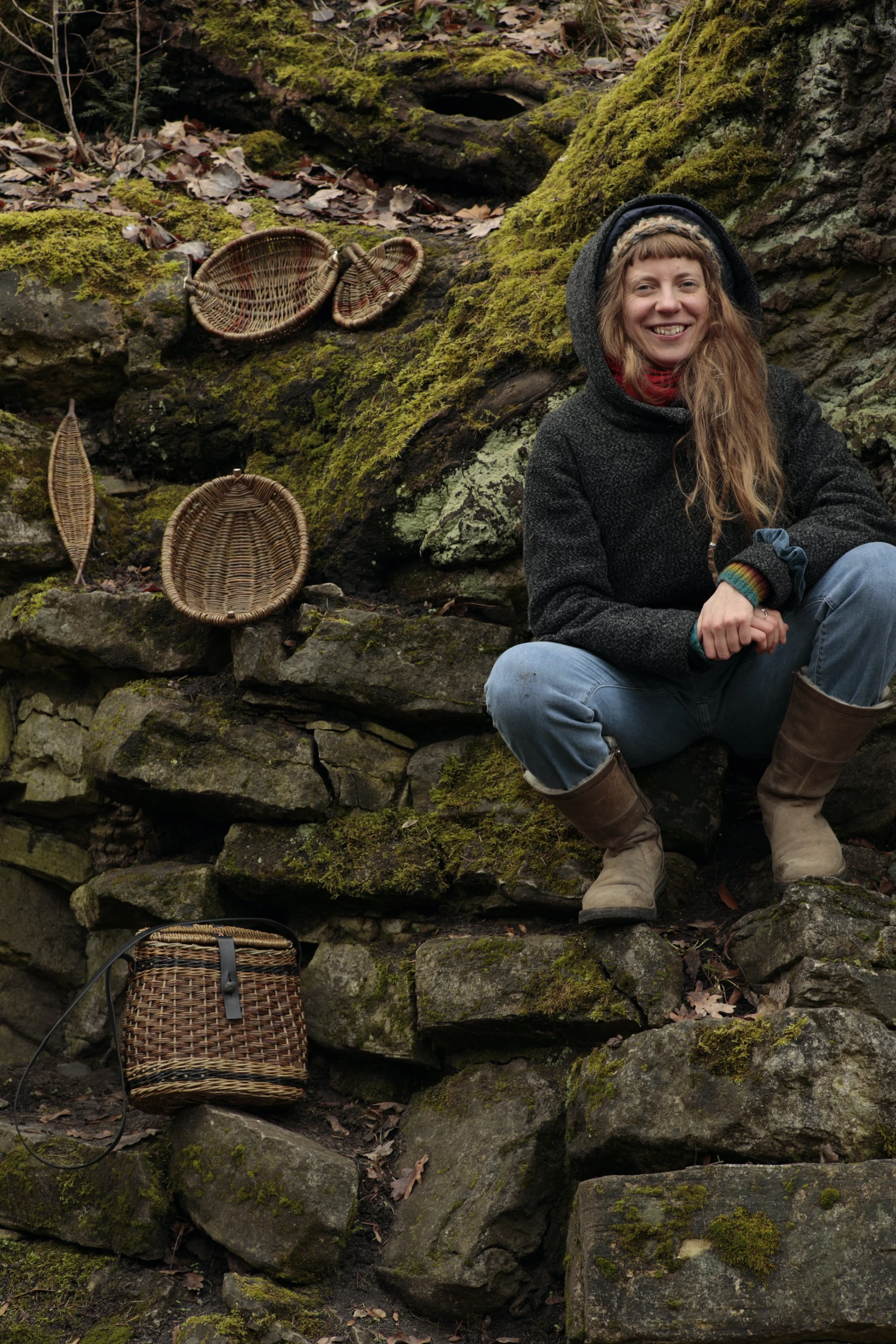 Persona sonriendo sentada en rocas cubiertas de musgo con cestas de mimbre alrededor en un entorno natural.